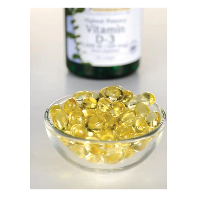 A small glass bowl filled with yellow Swanson Vitamin D3 5000 IU softgels, vital for bone health and a strong immune system, sits in the foreground, while a blurred bottle of the same stands behind.