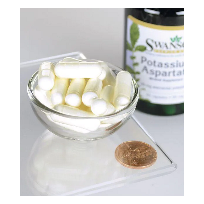 A Swanson bottle labeled "Potassium Aspartate 99 mg 60 Capsules" sits next to a small glass bowl filled with white capsules, emphasizing the supplement's role in maintaining electrolyte balance. A penny lies nearby on the reflective surface.