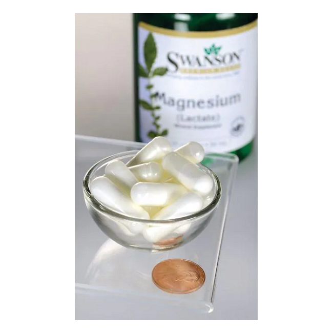 A bottle of Swanson Magnesium Lactate - 84 mg 120 capsules stands in the background, with a penny nearby and a bowl of white capsules on a glass surface, emphasizing its support for bone health.