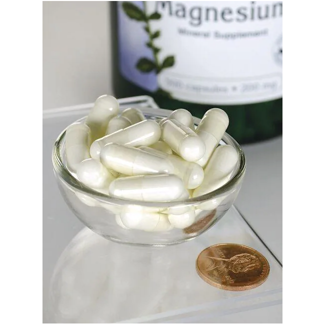 A small glass bowl holds white capsules next to a penny, showcasing their compact size. In the background, a bottle labeled "Swanson Magnesium Oxide 200 mg 500 Capsules" is partially visible, promoting bone health.