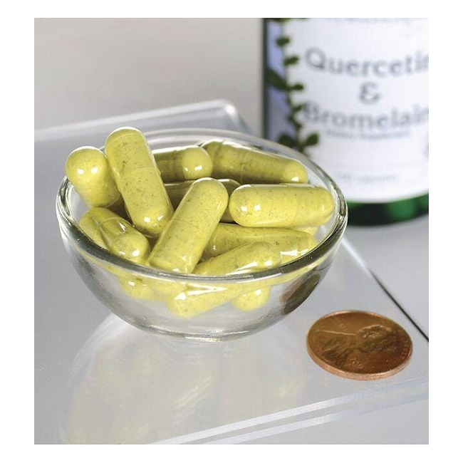 A small glass bowl filled with green capsules sits beside a coin for scale, emphasizing their compact size. In the background, a Swanson "Quercetin & Bromelain - Advanced Formula 100 Capsules" bottle peeks out, indicating their role in supporting immune health.