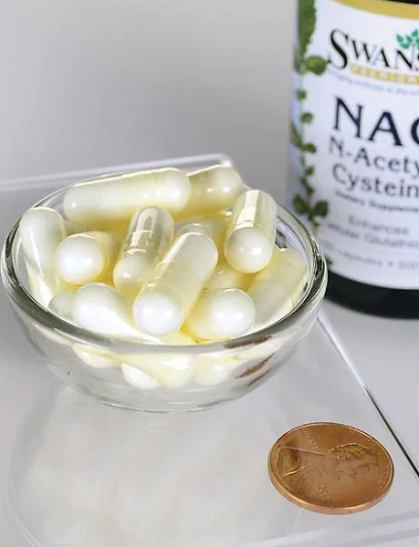 A small glass bowl contains clear capsules filled with white powder, alongside a copper penny for scale. Nearby, a partially visible bottle labeled "Swanson NAC - N-Acetyl Cysteine 600 mg 100 Capsules," known for its potent antioxidant properties and popularity as a dietary supplement, stands prominently.